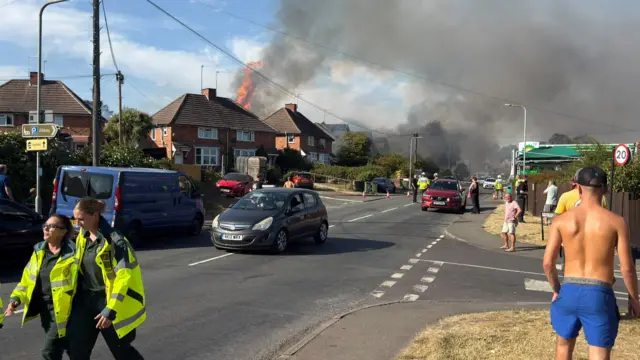Flames tower over a row of two storey houses while emergency services and members of the public stand in the road.