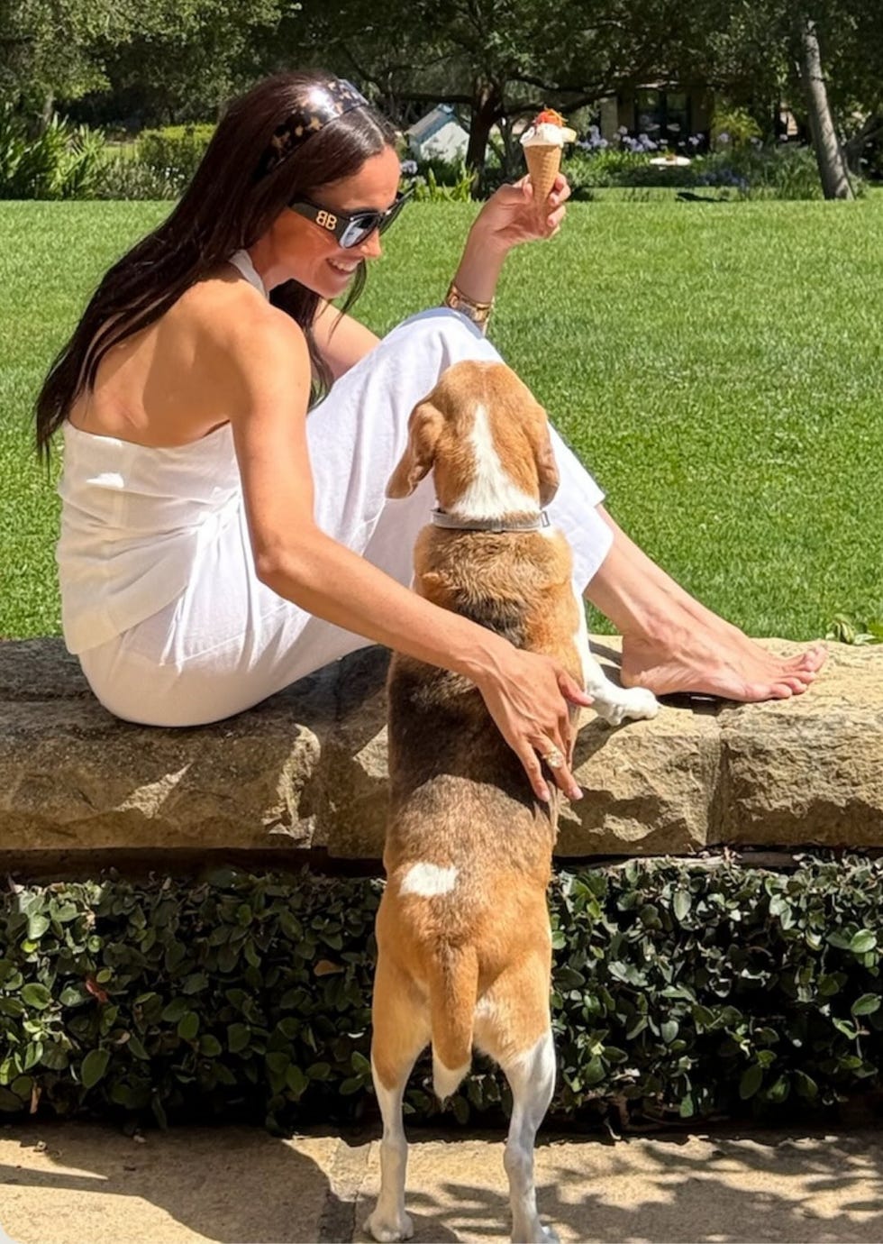 A woman in a white outfit enjoys ice cream while sitting on a stone wall with a dog beside her.