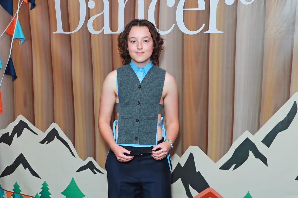 bella ramsey sunny dancer premiere person in a blue shirt and gray vest holding sunglasses, standing in front of a mountain themed backdrop.