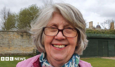 A woman standing outside on a grey day. She is smiling at the camera. She has short grey hair and is wearing glasses along with a pink jumper and a blue and green scarf.