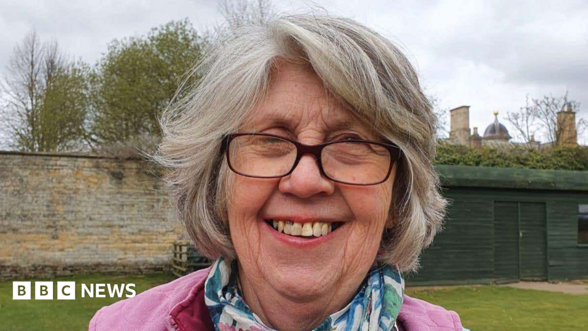 A woman standing outside on a grey day. She is smiling at the camera. She has short grey hair and is wearing glasses along with a pink jumper and a blue and green scarf.