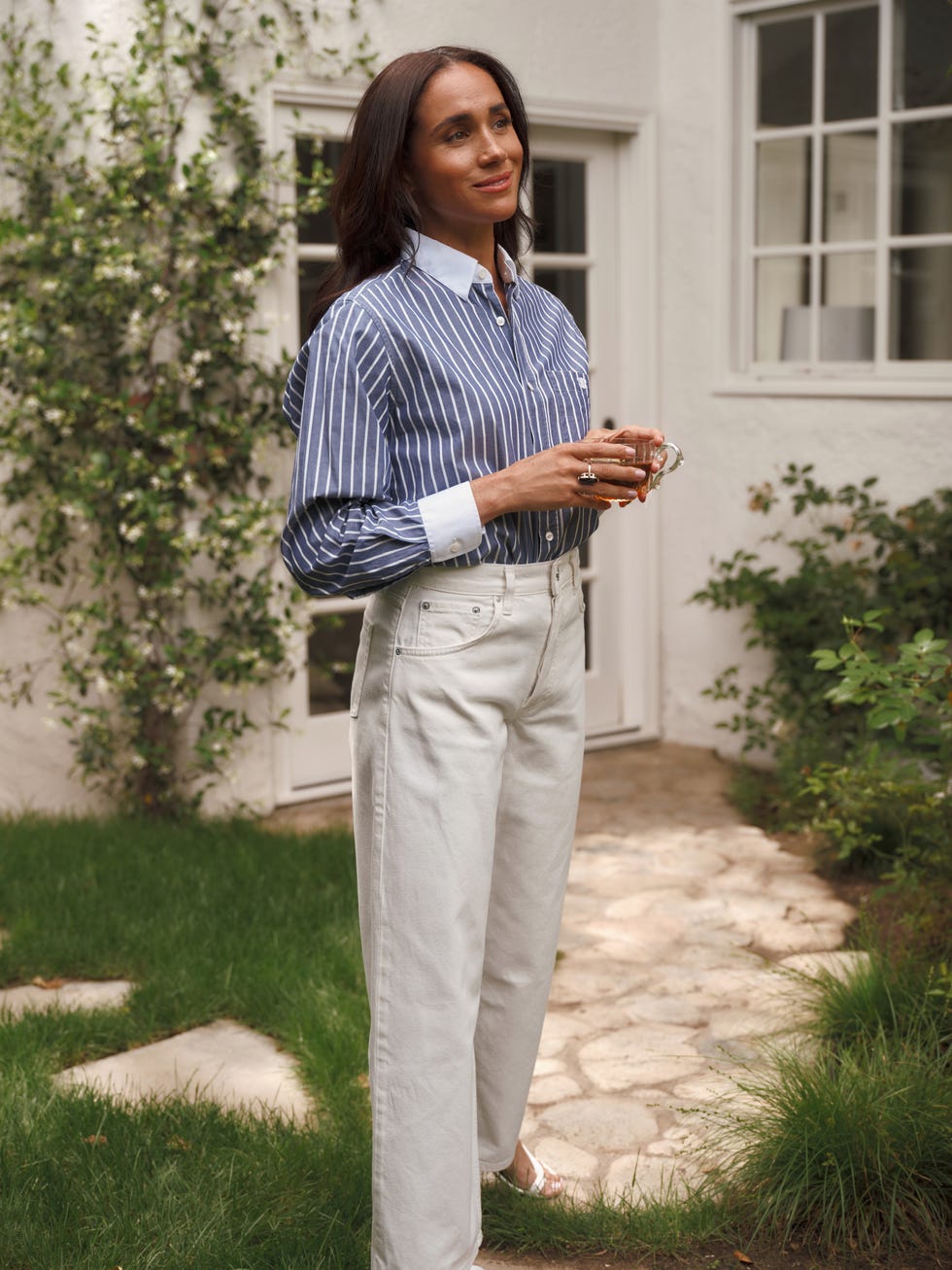 Person standing outdoors in a striped shirt and white pants, holding a drink with greenery in the background.