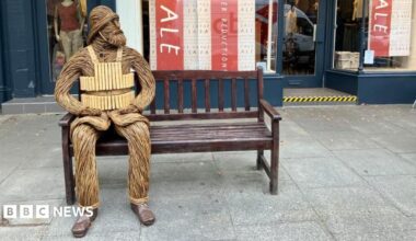 A wicker figure depicting a bearded RNLI crewman from 1886 - including a helmet and lifejacket - is sitting on a bench outside a shop in a town centre. The figure has its hands resting on its legs and is looking to its left.