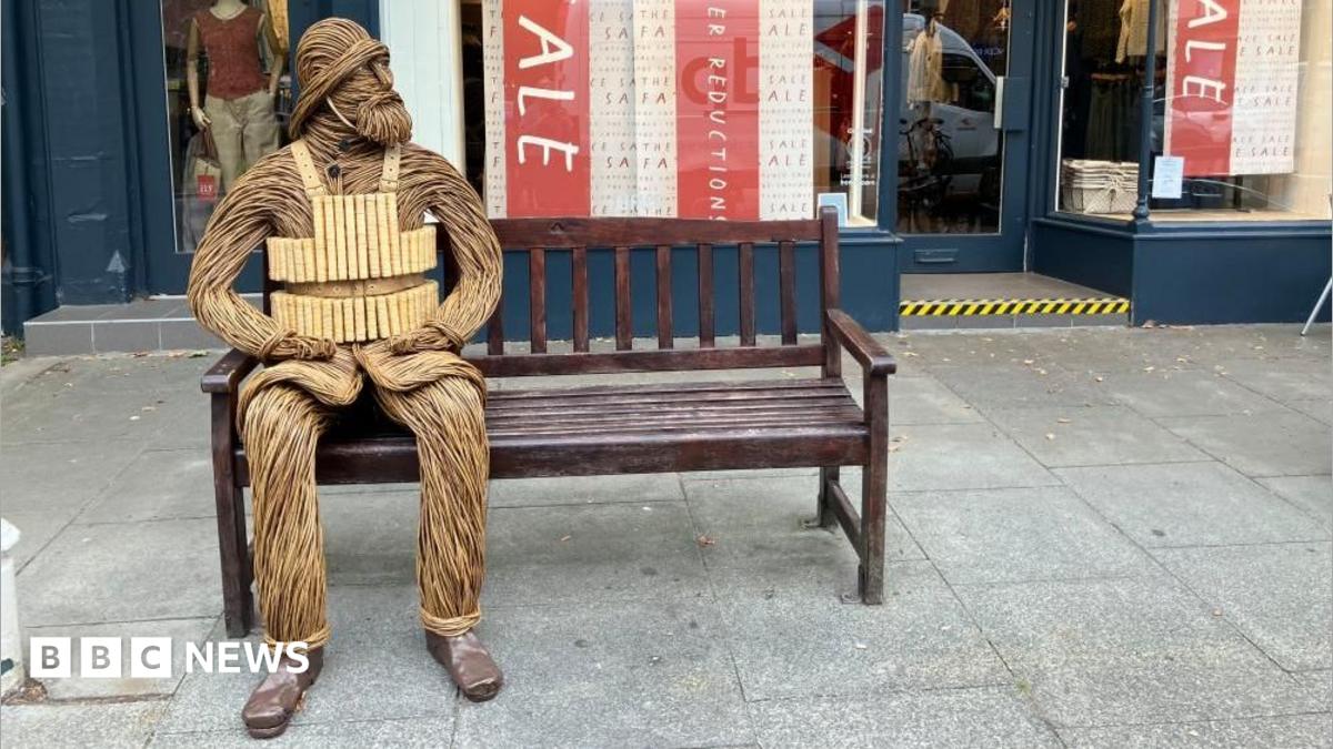A wicker figure depicting a bearded RNLI crewman from 1886 - including a helmet and lifejacket - is sitting on a bench outside a shop in a town centre. The figure has its hands resting on its legs and is looking to its left.