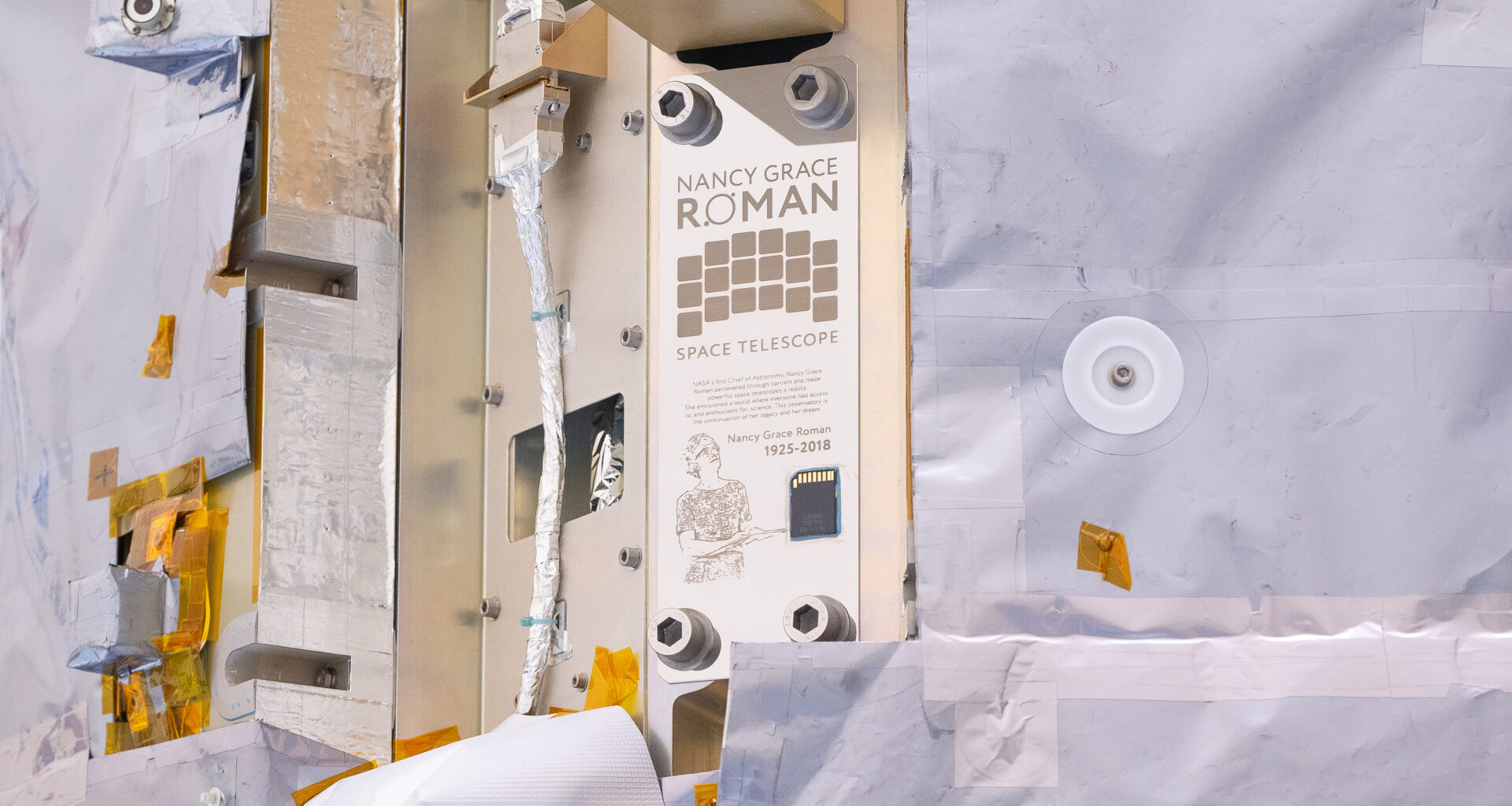 A close-up view of NASA's Nancy Grace Roman Space Telescope shows a commemorative plaque with writing on it, as well as an illustration of Roman, and a memory card.