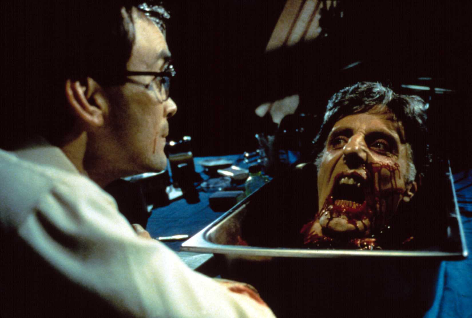 Lab-coated scientist (?) Herbert West (Jeffrey Combs) sits calmly as a blood-covered, severed, reanimated head screams at him from a basin on his lab counter in Re-Animator