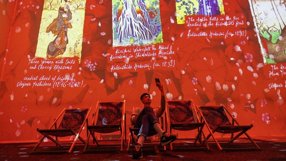 Person on phone in red-lit room with art projected on walls and deck chairs at Van Gogh: The Immersive Experience in Cincinnati, US.