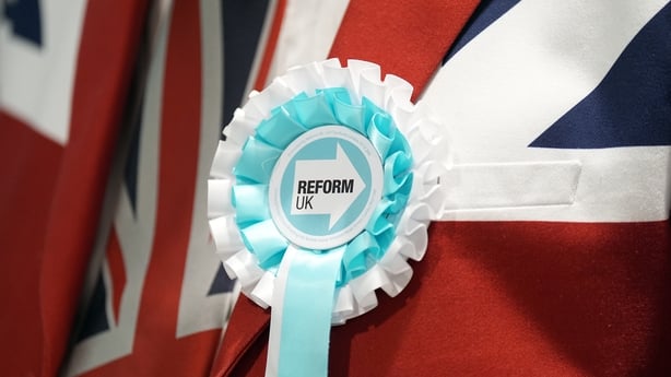 A man in a union jack blazer wears a Reform UK rosette