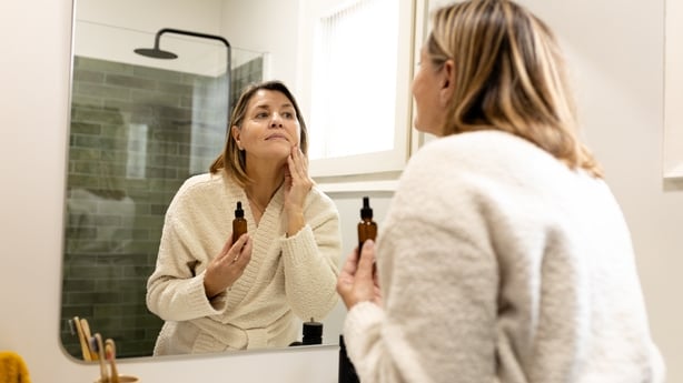 Woman wearing bathrobe looking in the mirror and applying moisturizer on her face in the bathroom