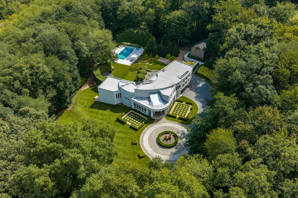 Modern house surrounded by lush greenery, featuring a circular driveway, manicured lawns, and a swimming pool.