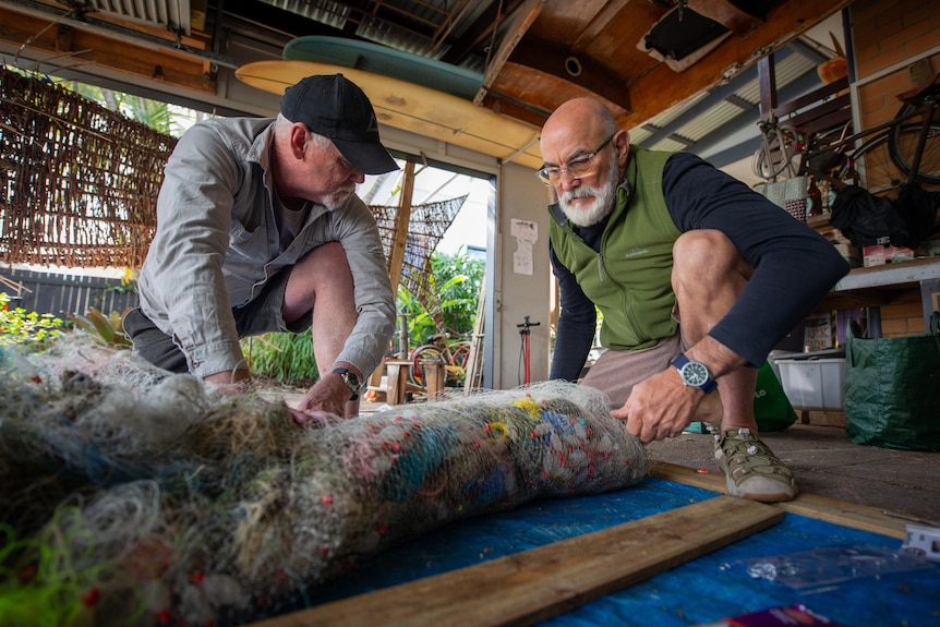Two men kneel on the floor of a workshop rolling fishing line and discarded plastics into a sausage-shaped net.