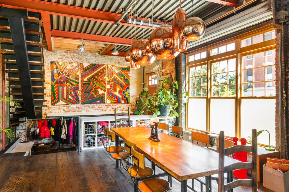 A bright, stylish dining area featuring a long wooden table, colorful artwork, and large windows.