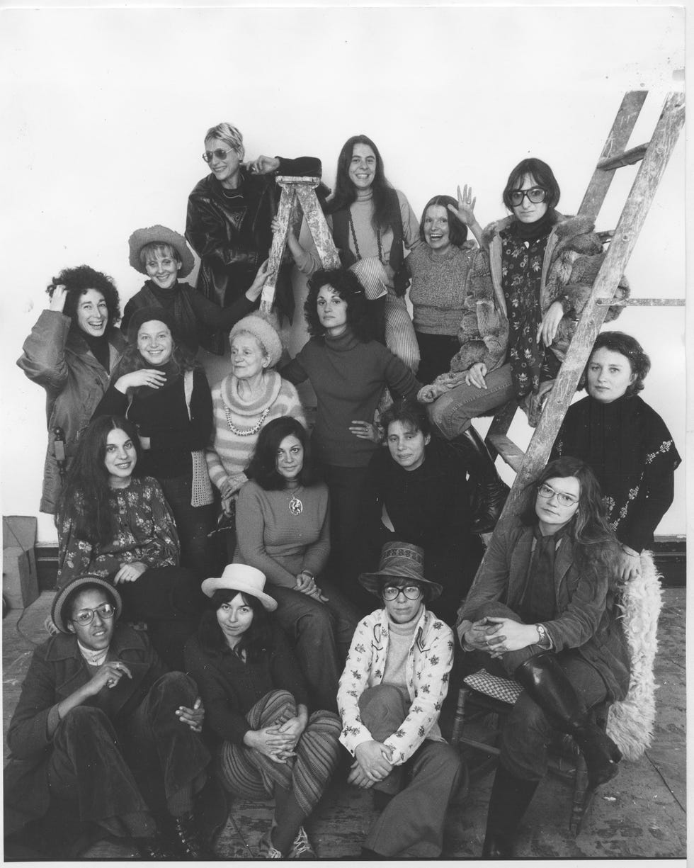 A diverse group of women posing together, some sitting on a ladder and others on the floor, in a studio setting.