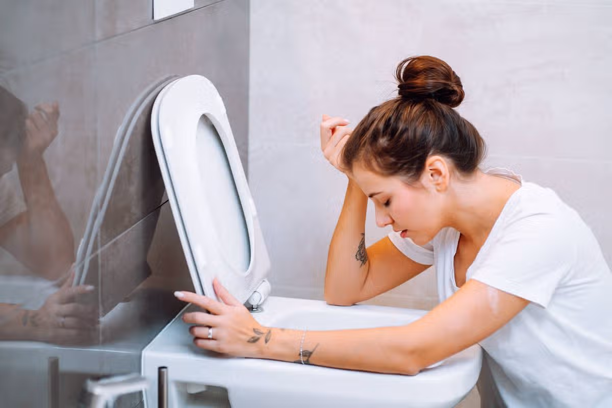 A woman in a white t-shirt is seated on a toilet bowl with her head resting on her left hand, appearing distressed, while the toilet seat is open.