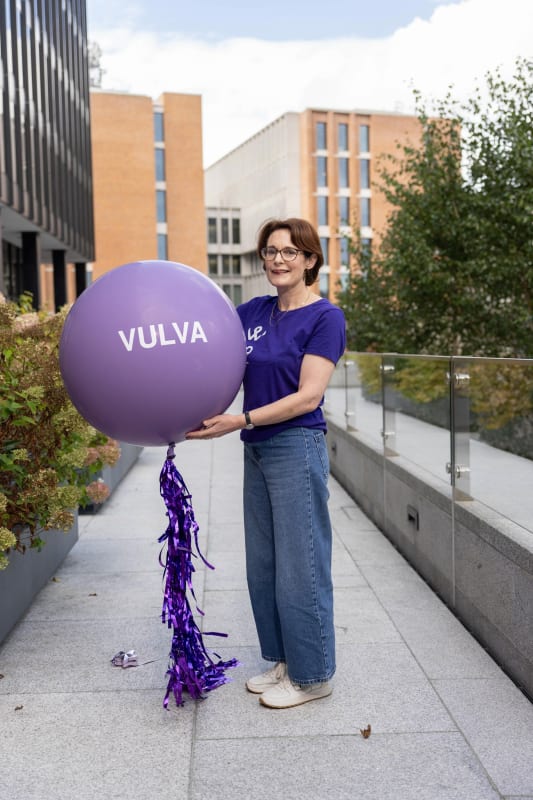 Mairead McMahon holding a large purple balloon printed with the word VULVA, standing on an outdoor terrace.