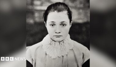 A black and white photograph of a young woman with hair tied back, looking straight at the viewer. She is wearing a white, frilly ruffle over a light-coloured top.
