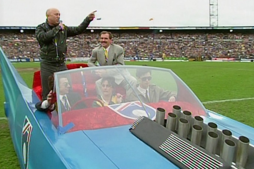 A bald man in a black leather jacket stands in the red seat of a light blue open air car pointing to a crowd in a stadium.