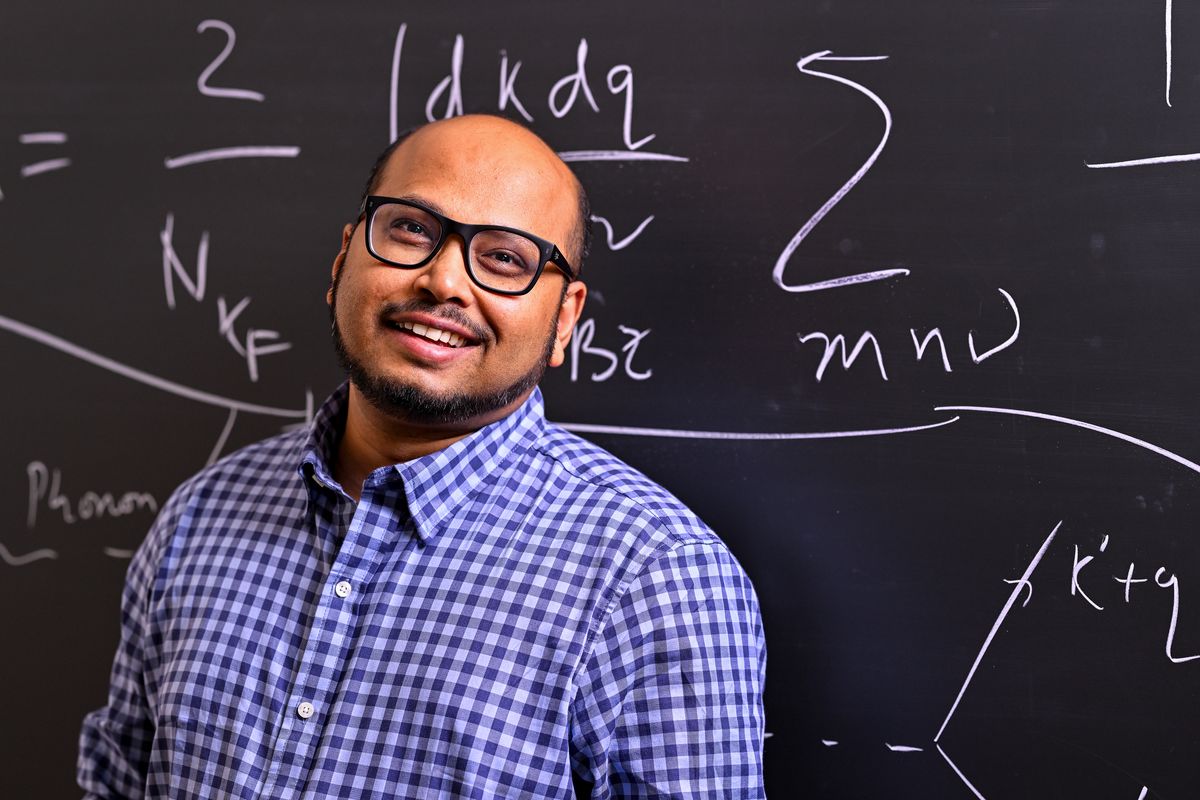 A man with glasses smiles in front of a chalkboard
