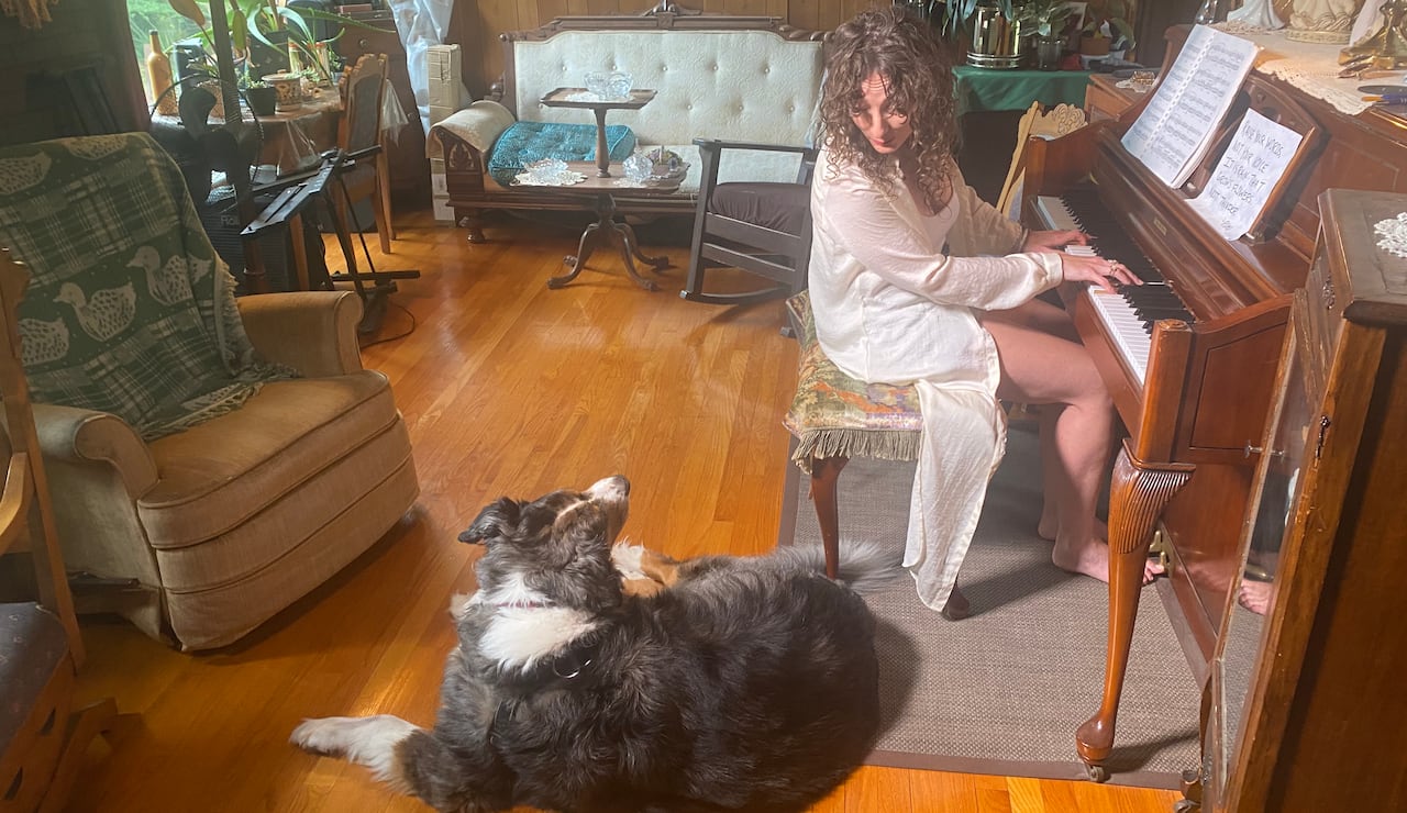 A woman with long curly brown hair plays a piano while looking down at a large dog looking back up at her.