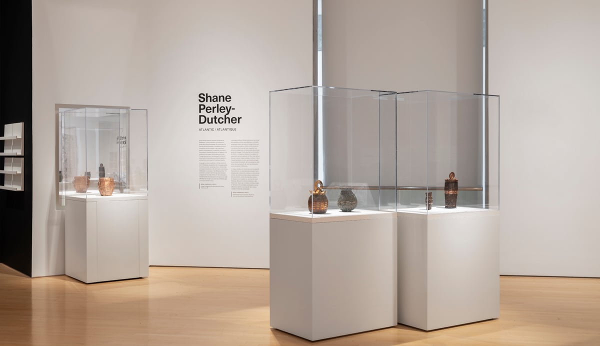 a large white space, with text on wall reading Shane Perley-Dutcher, and glass cases with metal woven baskets on display.