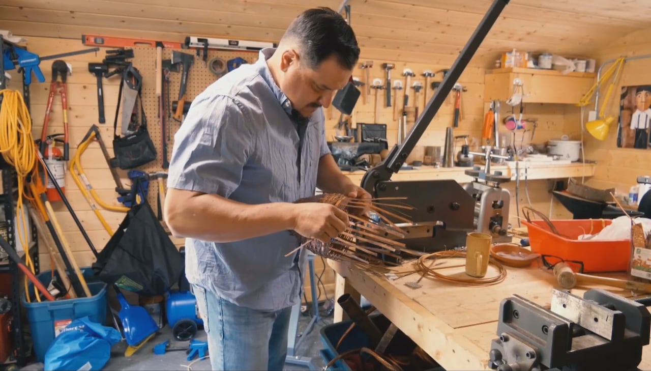 A man working on weaving a basket with metal strips, in a workshop surrounded by tools.