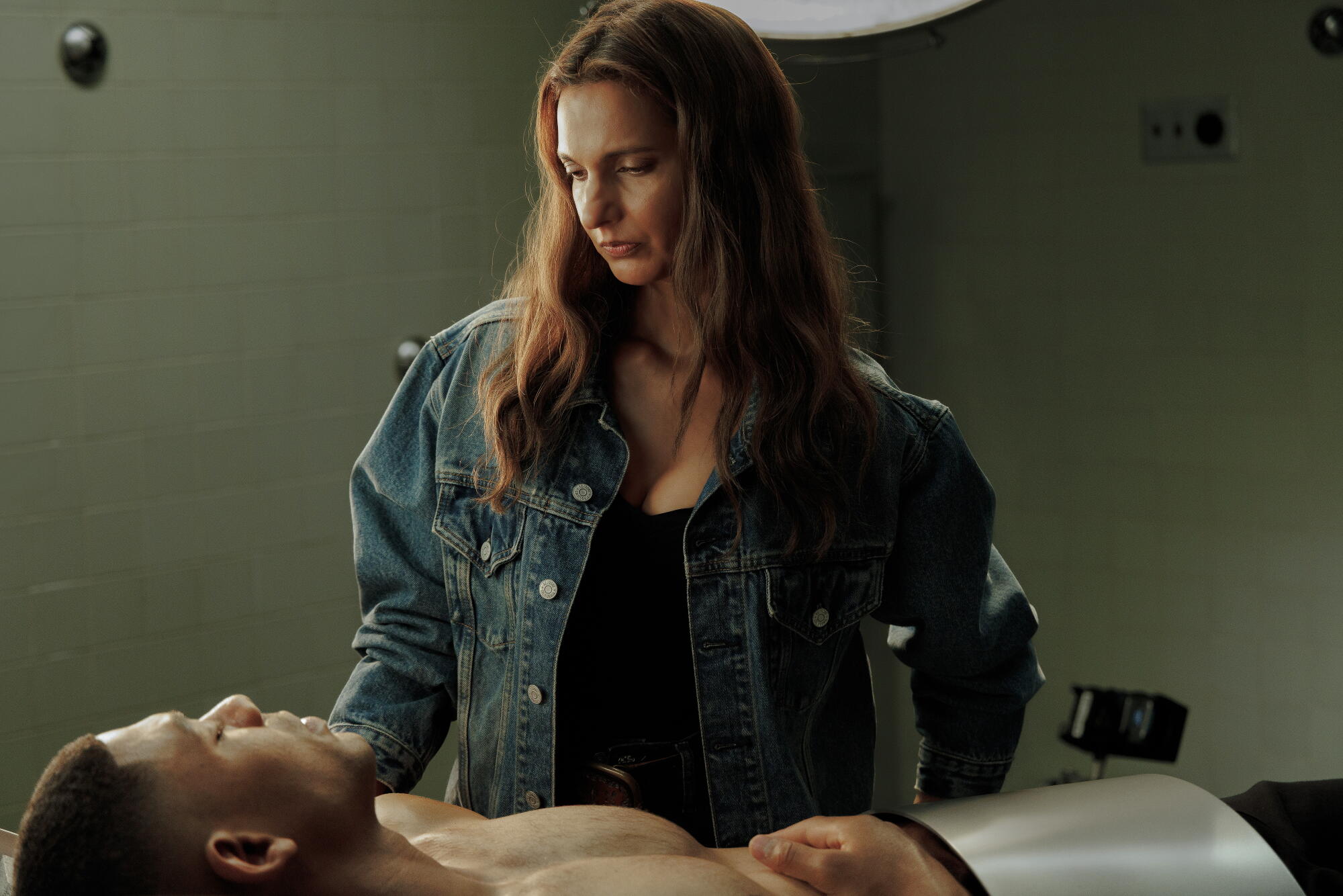 A woman with long brown hair in a denim jacket and black T-shirt looks down at an injured shirtless man laying on a table.