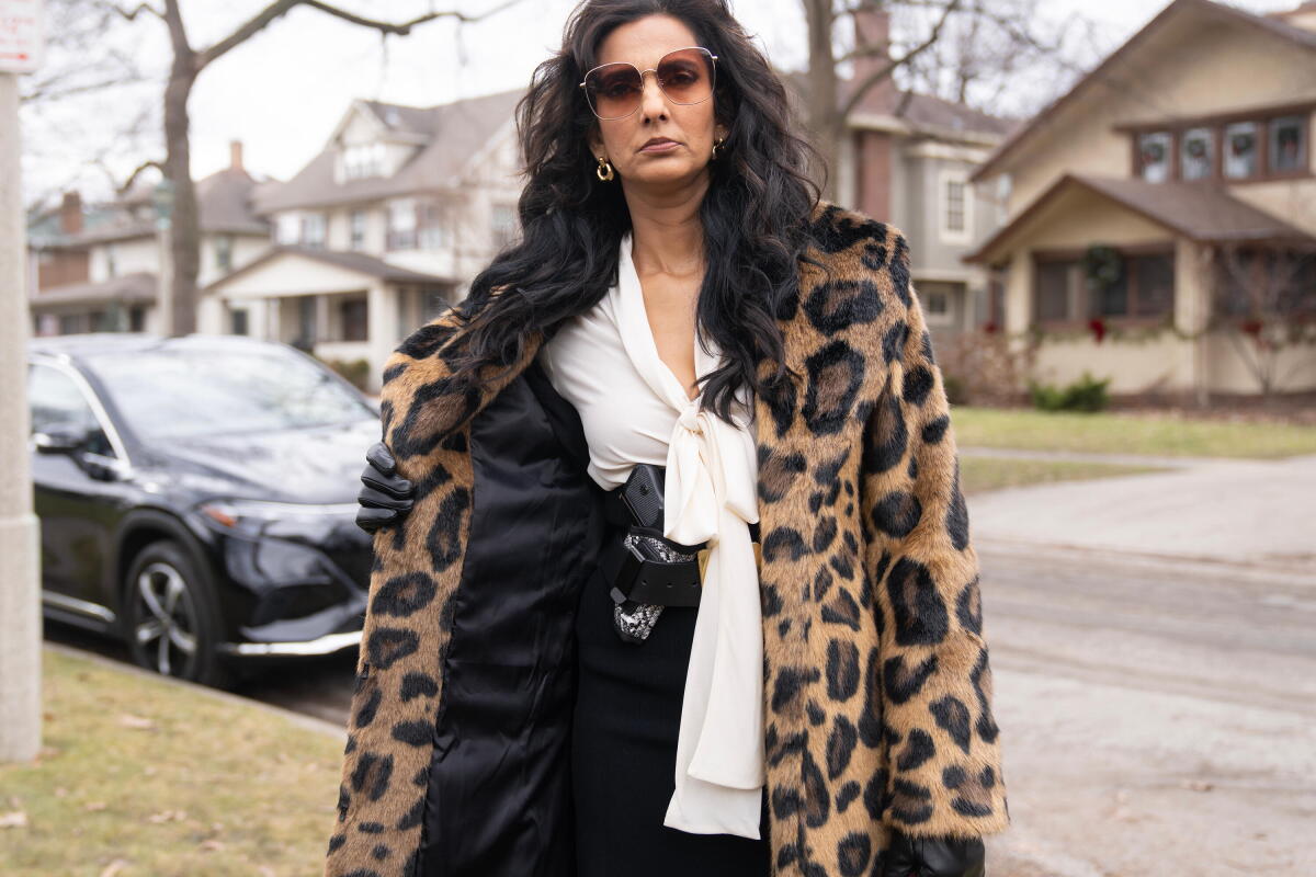 A woman in sunglasses, a leopard print fur coat, white shirt and dark slacks flashes a gun in holster at her waist.