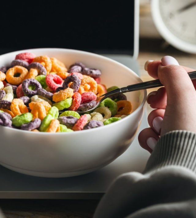 Woman eating colored breakfast cereal in front of laptop, workplace breakfast, quick healthy breakfast in office in the morning.