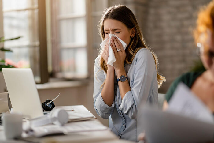 A woman sneezes into a tissue at her desk, depicting seemingly innocent symptoms that turned into serious issues.