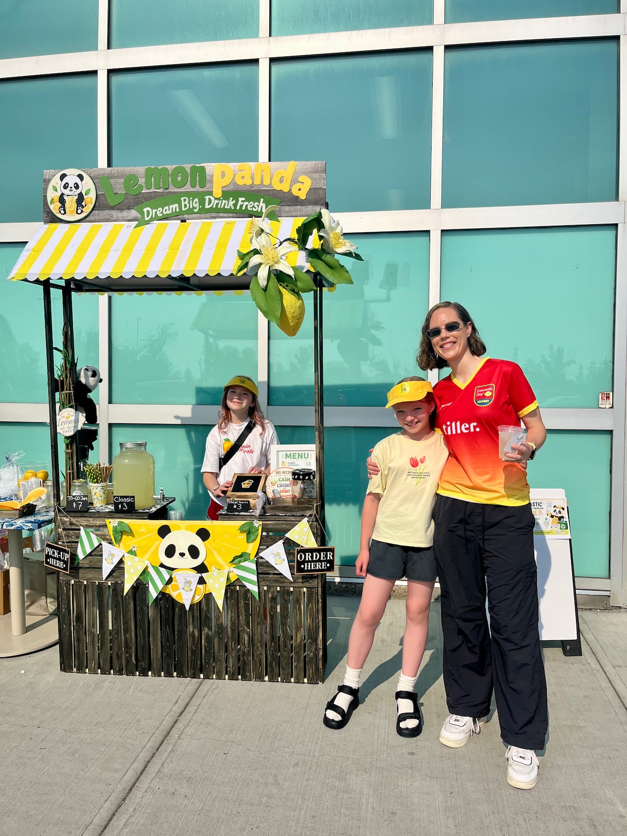 A woman smiles next to two kids selling lemonade.