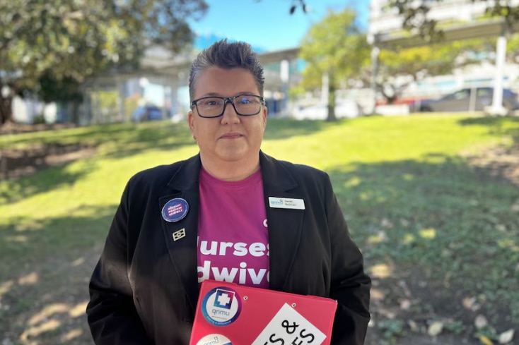 A woman with glasses holds a red folder in a park.
