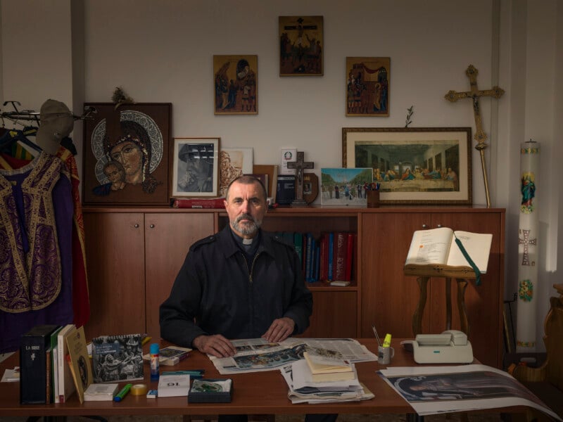 A middle-aged priest with a beard sits at a desk in his office, which is decorated with religious icons, a crucifix, and vestments.