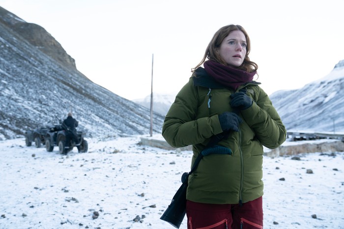Rose Leslie as Kirsten Longacre in Vigil, wearing a coat and scarf, stood in a snowy, mountainous landscape and with a shotgun strapped to her back.