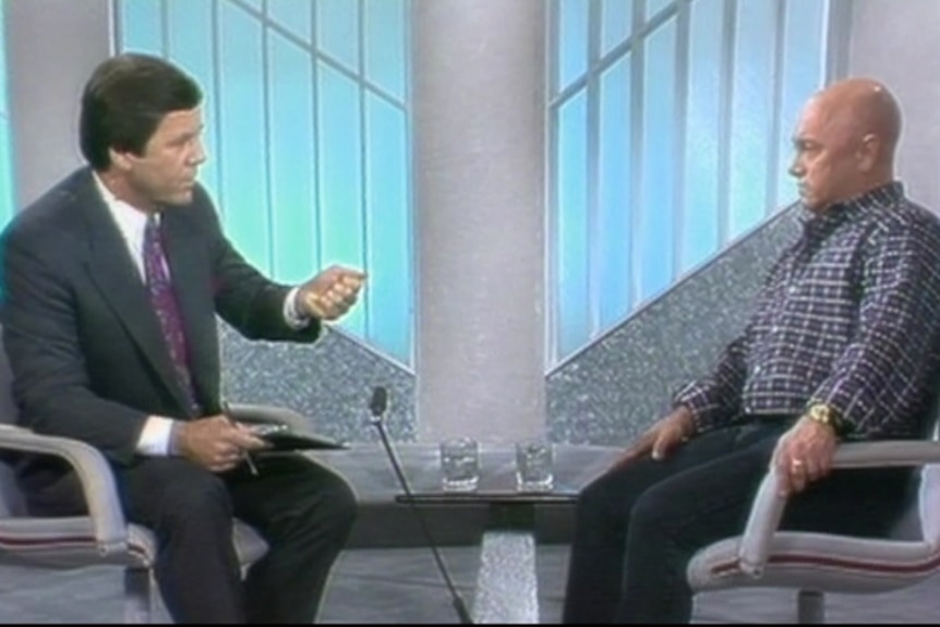 A dark haired man in a suit and a bald man in a checked shirt sit opposite each other in a tv studio.