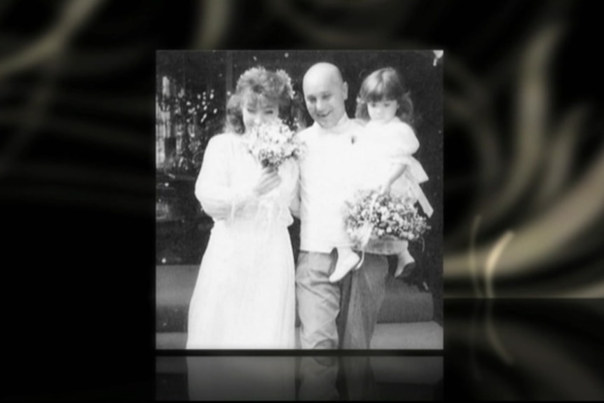 A black and white photo of a woman in a wedding dress with flowers in front of her face and a bald man holding a young girl.