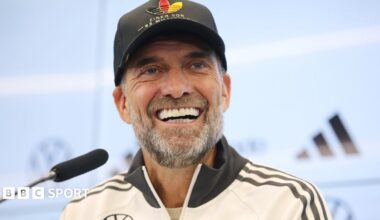 Germany boss Jurgen Klopp, wearing a white jacket and black baseball cap, smiles during a news conference with a microphone in front of him