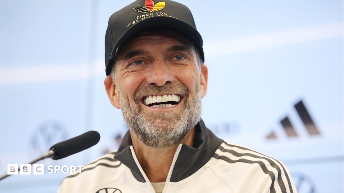 Germany boss Jurgen Klopp, wearing a white jacket and black baseball cap, smiles during a news conference with a microphone in front of him