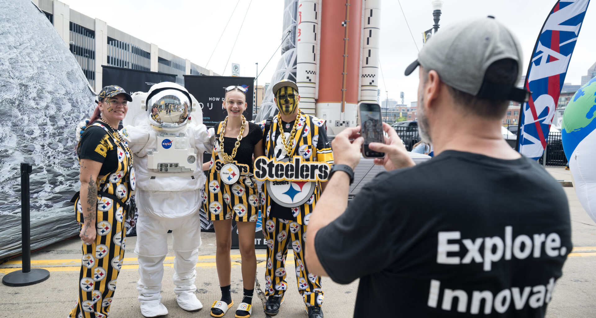 Three Pittsburgh Steelers fans, dressed in black and yellow clothes with the Steelers' logo on it, pose with an astronaut in a spacesuit. The reflective visor is closed. In the foreground, a man takes the group's picture with his phone.