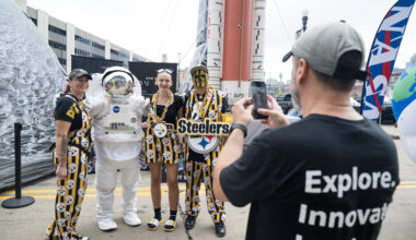 Three Pittsburgh Steelers fans, dressed in black and yellow clothes with the Steelers' logo on it, pose with an astronaut in a spacesuit. The reflective visor is closed. In the foreground, a man takes the group's picture with his phone.