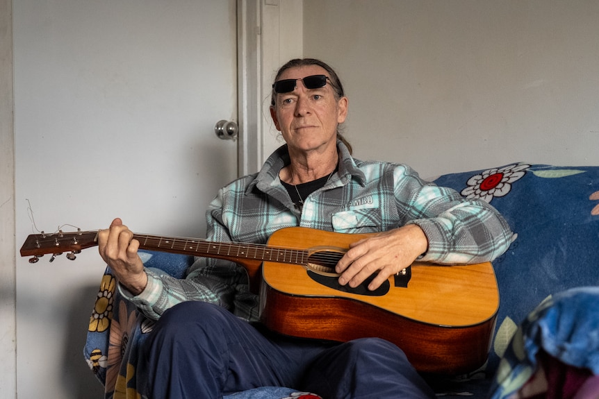 A man wearing a checkered shirt is sitting and playing a guitar.