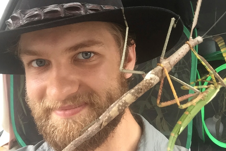 A smiling, bearded man in a dark, broad-brimmed hat holds a stick with a large insect clinging to it.