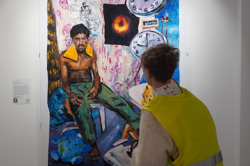A woman in a high-viz vest looks at a painting depicting a shirtless man sitting on a chair in a room with clocks