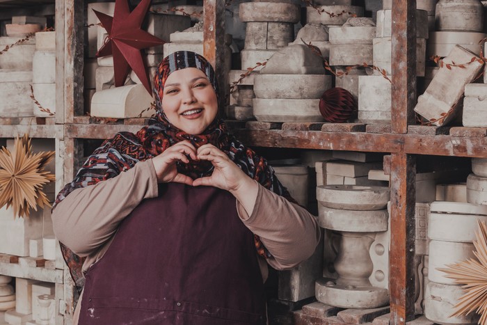 Fatiha El Ghorri stood in a pottery studio smiling ahead as she makes a heart shape with her hands.