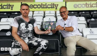 Dale Vince and Dr Simon Opher sit in a stand at Forest Green Rovers' stadium holding a flyer advertising the scheme. Vince is wearing a black and white T-shirt and black and white trousers and he looks quite serious. Opher is wearing a white shirt with the sleeves rolled up and beige trousers and he is smiling.