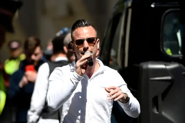 Danny Tommo seen wearing a white shirt and sunglasses, with his hair slicked back. He's speaking into a microphone held close to his mouth