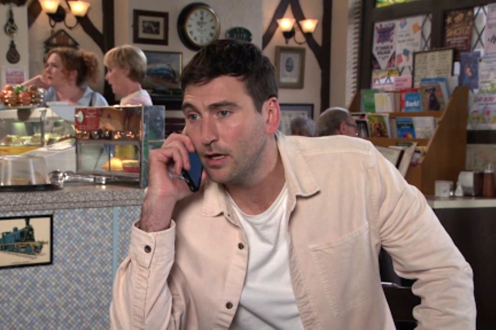 coronation street kit green man talking on phone at a busy café counter.
