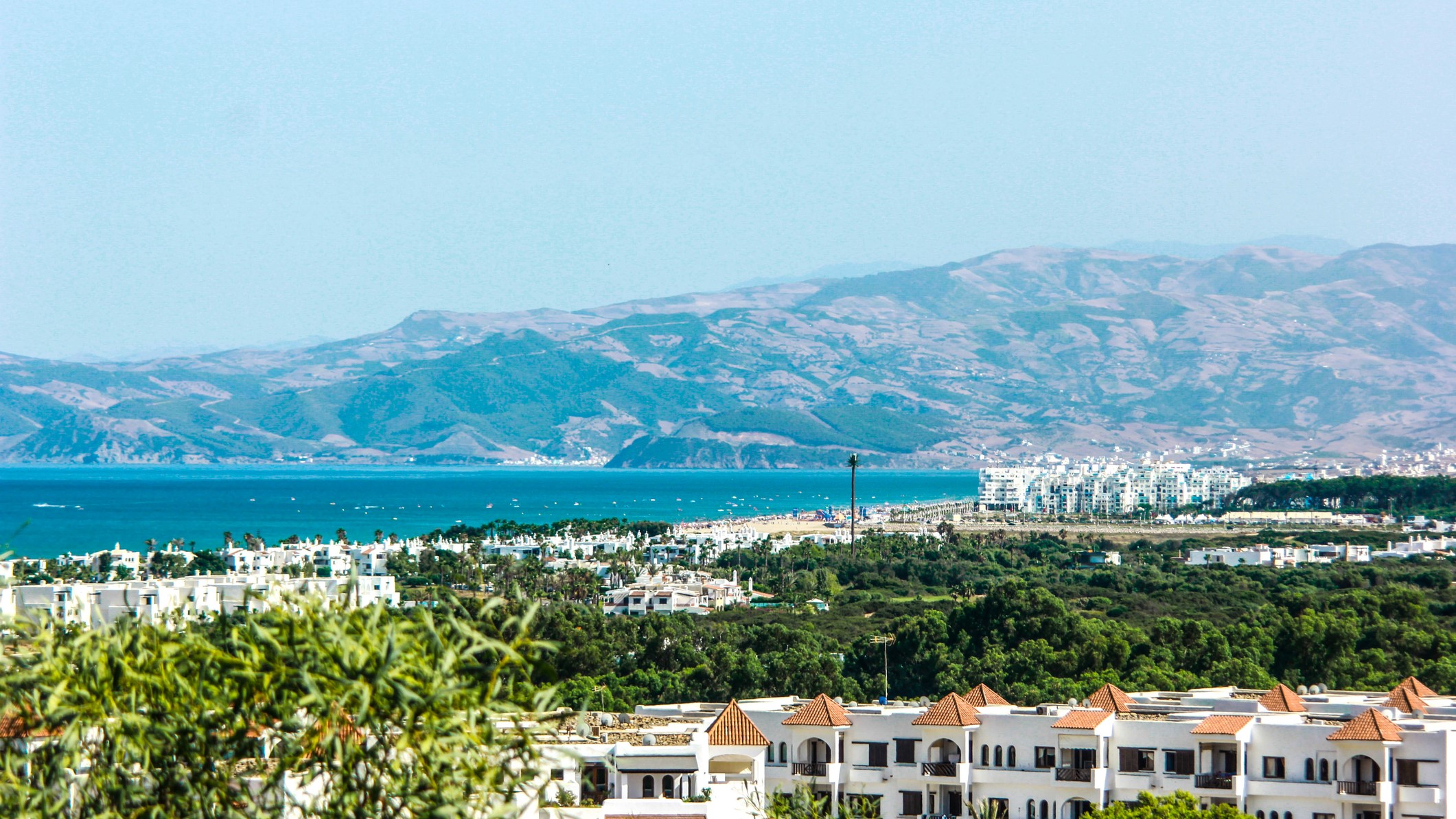 Martil beach, Morroco. White buildings and trees in the foreground with a beach scene in the middle of the image and then large hills and mountains in the background.
