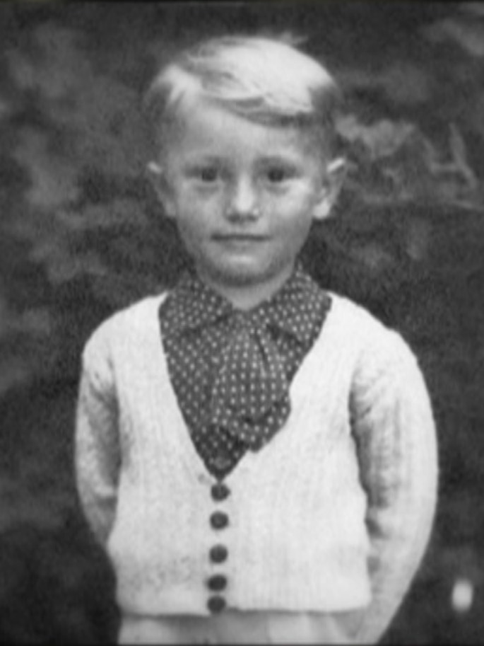 A black and white photo of a light haired boy in a white cardigan over a polka dot collared shirt.