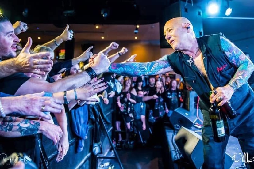 A crowd presses against a barrier reaching out to a bald tattooed man on stage who reaches to them.