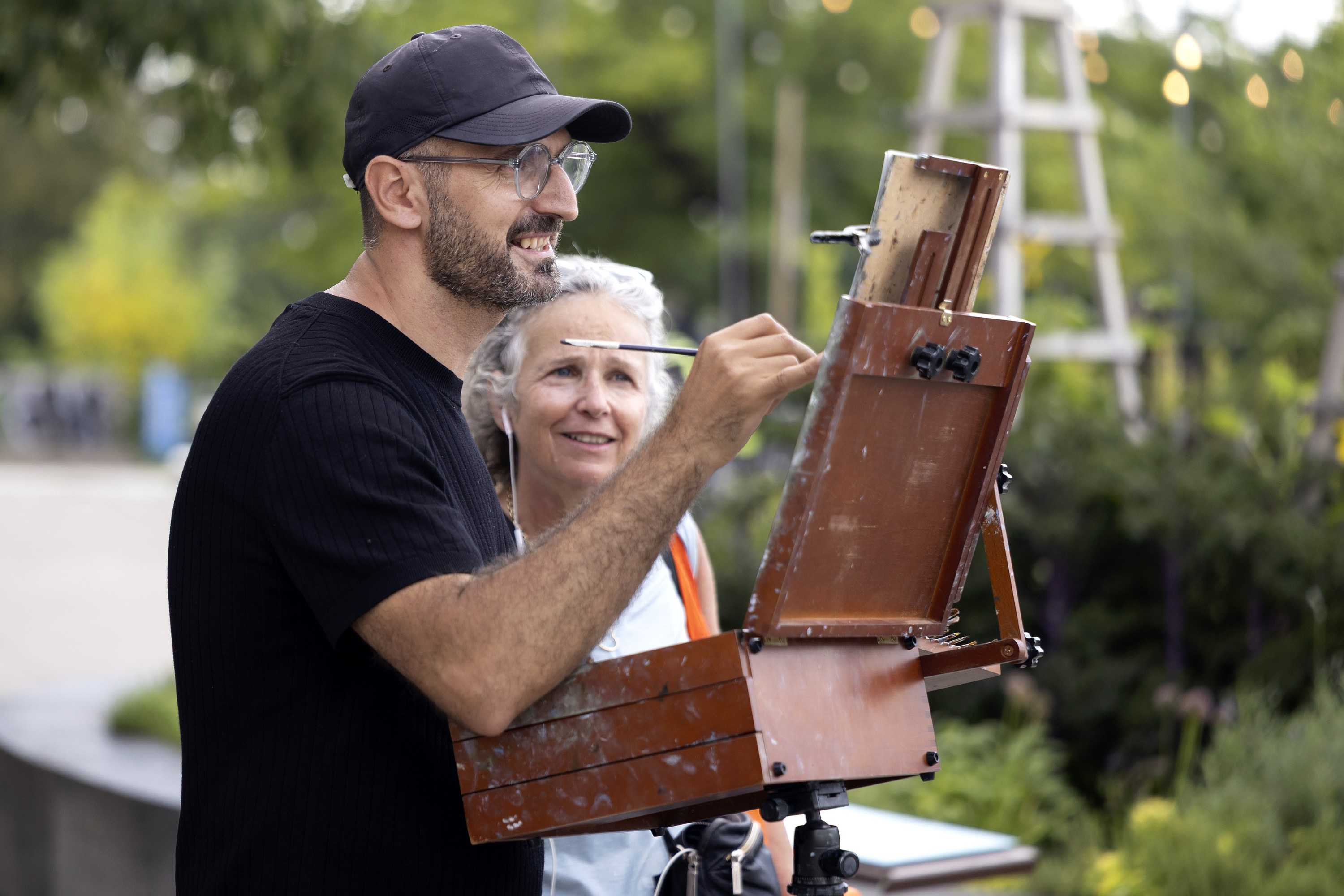 Man paints at easel outdoors while woman standing beside him watches 
